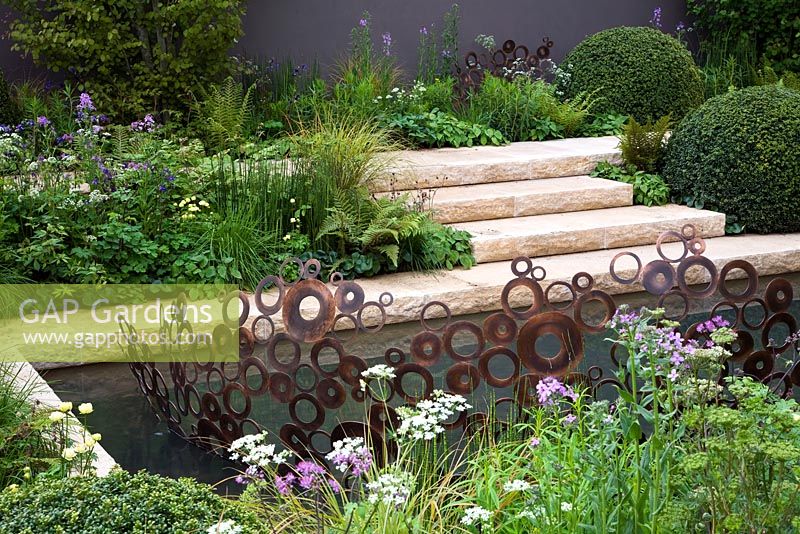 A garden inspired by the Arts And Crafts Movement. Formal paths and terraces combine with a water channel. Focal point is an energy wave sculpture made from copper rings. Planting style is woodland edge with buxus domes - The M & G Garden