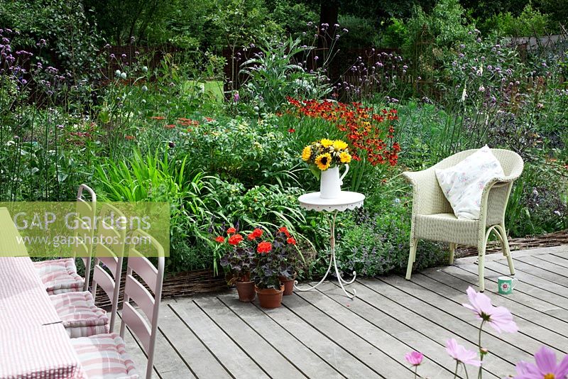 Lloyd Loom chair with cushion, small table with vase of Sunflowers in decked garden with densly planted border. Pots of Pelargoniums. Verbena bonariensis, Heleniums, Achillea, Nepeta, Rose bush with hips and Cardoon.
