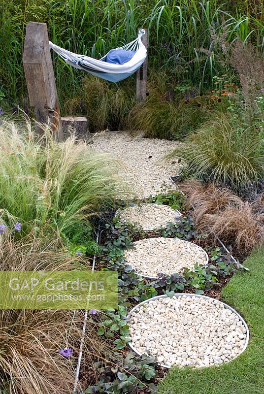 Hammock in small gravel area surrounded by ornamental grasses and path of circular containers filled with gravel, bark chippings and Viola in the 'One Man Went To Mow' garden  RHS Tatton Flower Show 2012