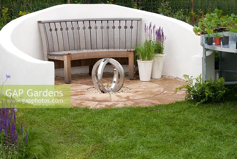 Wooden curved bench on circular patio with central sculpture and white rendered wall in the 'Ring the Changes' garden 