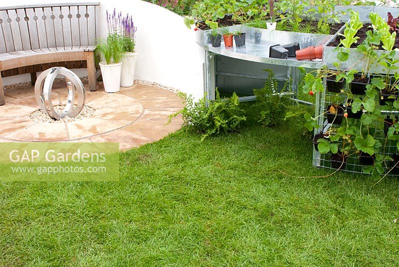 Wooden curved bench on circular patio with adjacent aluminium staging for potting and growing vegetables and strawberries in the 'Ring the Changes' garden, RHS Tatton Flower Show 2012