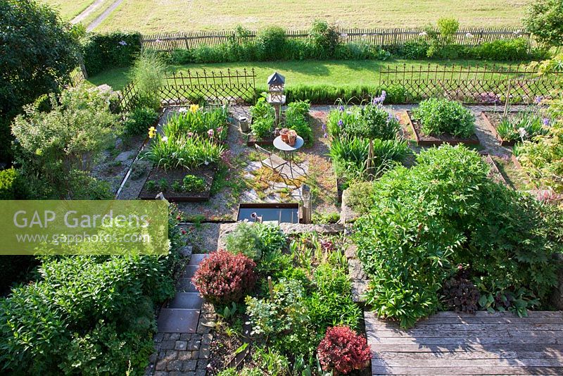 View from above onto a modern structured country garden with a small rest area, vegetable patches and a wooden terrace. Plants include Berberis thunbergibi 'Atropurpurea', Dictamnus albus and Paeonia
