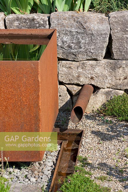 Next to corten steel water basin, a pipe coming out of a dry stone wall brings water to a rill