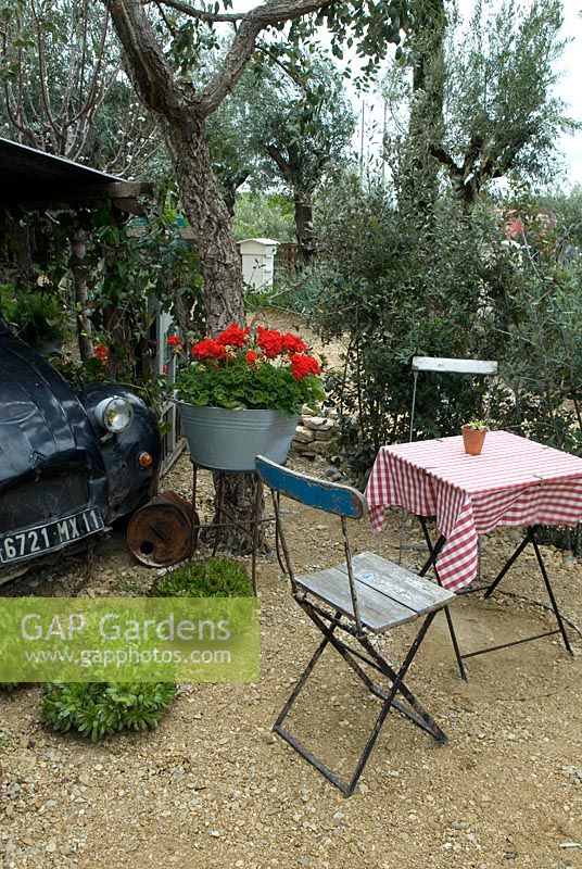 Table and chairs in garden of rural French cafe, with Olive trees, red Geraniums in galvanised bath and old Citroen car  - 'Reposer Vos Roues/Rest your Wheels' Show Garden, Gold Award, Malvern Spring Show 2013