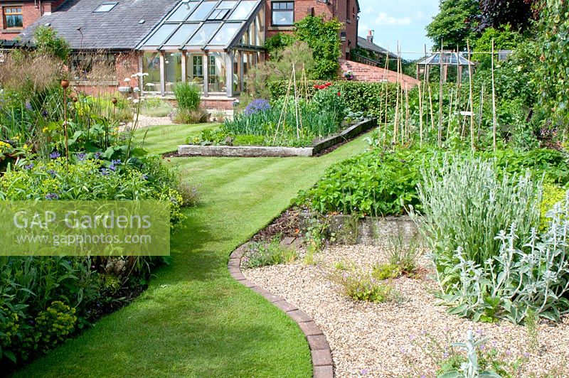 Curving lawn edged with block paving, herbaceous border, graveled area and raised vegetable beds in cottage garden in June