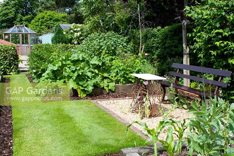 Graveled seating area with bench and raised vegetable beds in cottage garden surrounded by mature trees
