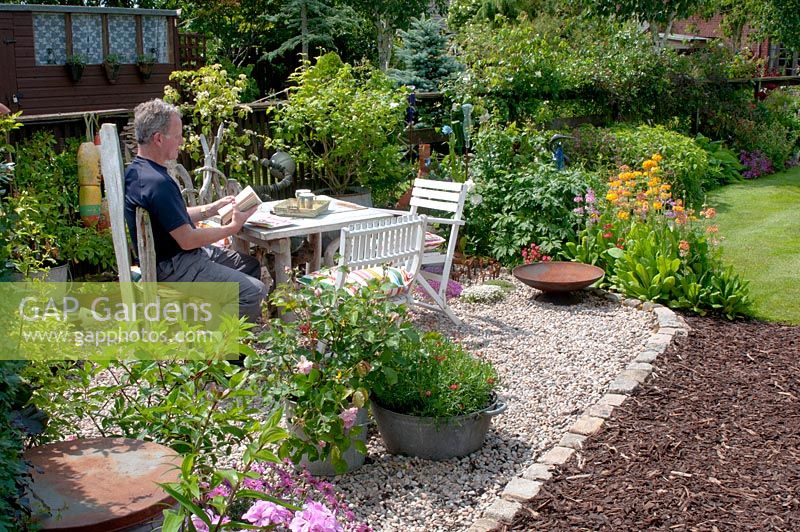 Man seating in graveled corner of cottage garden with small table and chairs and collection of vintage galvanised containers - planting includes; Rosa, Primula, Argyranthemum and Geranium 