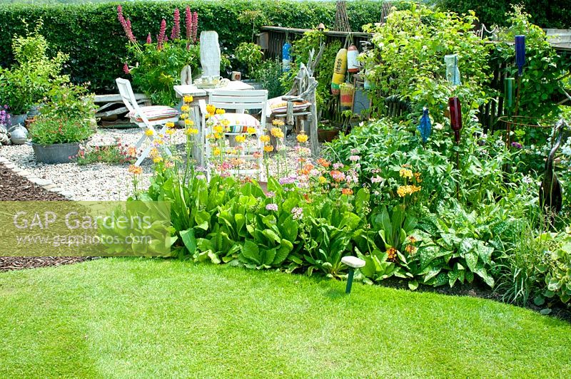 Seating area with small square table and chairs made of drift wood on graveled corner of cottage garden with collection of vintage galvanised containers fishing floats and planting including Lupinus, Primula and Geranium 