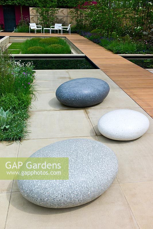 Large sculptural stone pebbles on paved area with borders of mixed perennials, a sunken area, pools of water and a wooden path in The Brewin Dolphin Garden 