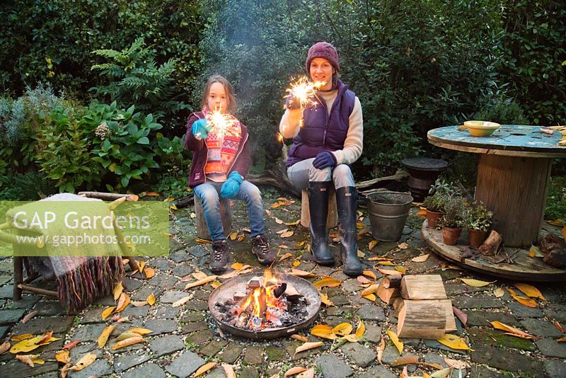 Mother and daughter lighting sparklers by a firepit in an autumnal back garden.