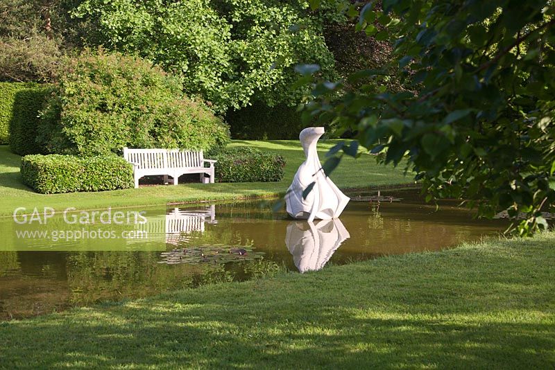 Hokusai's Boat sculpture in tranquil pond garden with clipped hedging and wooden seat.  Farleigh House, Hampshire
