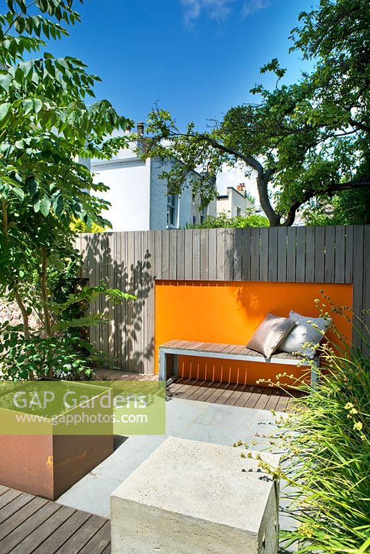 Modern contemporary garden in Brighton with decking, orange panels on walls, Metal water feature, wooden bench, Ophiopogon and Aralia. 