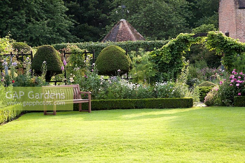 Clipped topiary and garden seat overlooking formal lawn with summer planting of Rosa, Papaver, Leucanthemum, Digitalis, Euphorbia, Fennel. Chenies Manor, Buckinghamshire
