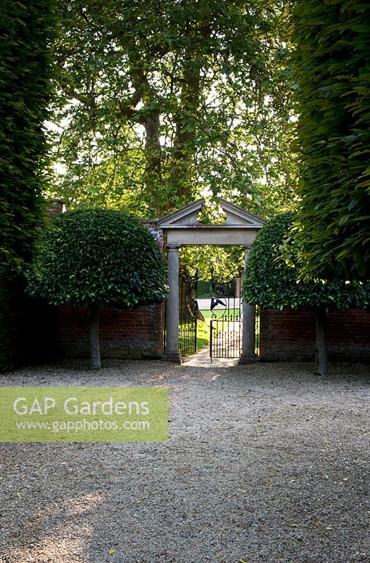 View from French-style pleached allee to formal gate in walled garden with ornate stone pediment and twin symmetrical topiary trees - Seend, Wiltshire