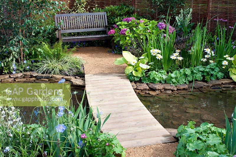Wooden bridge over pool leading to seating area with bench and colourful beds in the 'Ripples on The Water' garden designed by Muddy Boots -  Southport Flower Show 2013