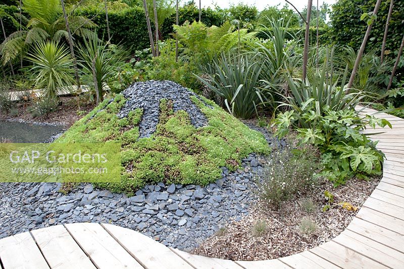 Title: Pour L Amour de Tongariro. Garden with wooden path passing several volcanos planted with sedums and covered with broken black slates.