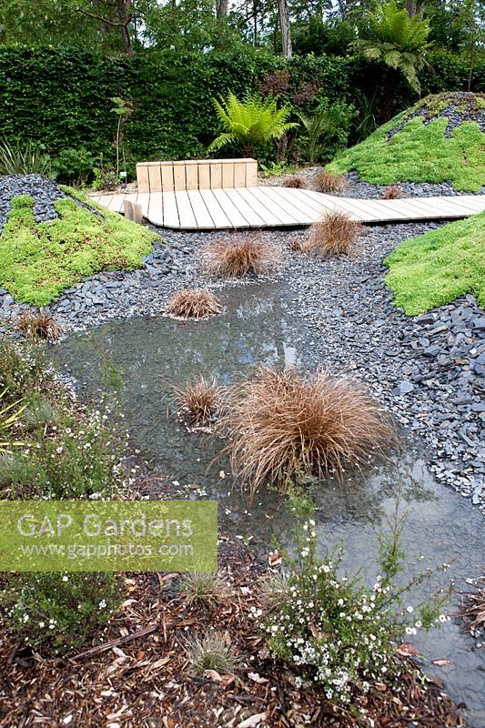 Title: Pour L Amour de Tongariro. Garden with wooden path passing several vulcanos planted with sedums and covered with broken black slates.