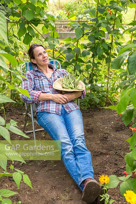 Woman relaxing in the Bean house with a harvest of Runner Bean 'Wisley Magic'. 