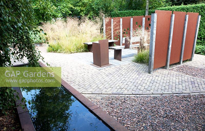 Pond with rusted metal edging in front of a relaxing garden area with rusted screens 