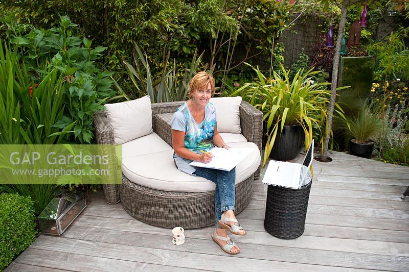 Garden designer, Angie Barker working on ratta sofa on Ipe decking surrounded by planting of Phormium 'Yellow Wave', Crocosmia 'Lucifer' and Phyllostachys nigra. A full standard Photinia fraseri 'Red Robin' provides privacy