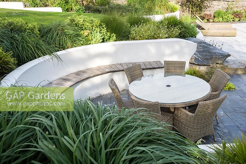 Contemporary terrace with curving rendered white retaining wall, inset wooden bench and rattan garden furniture. 