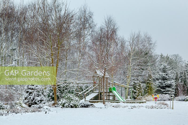 Country garden in winter with childrens play area and deciduous forest behind 