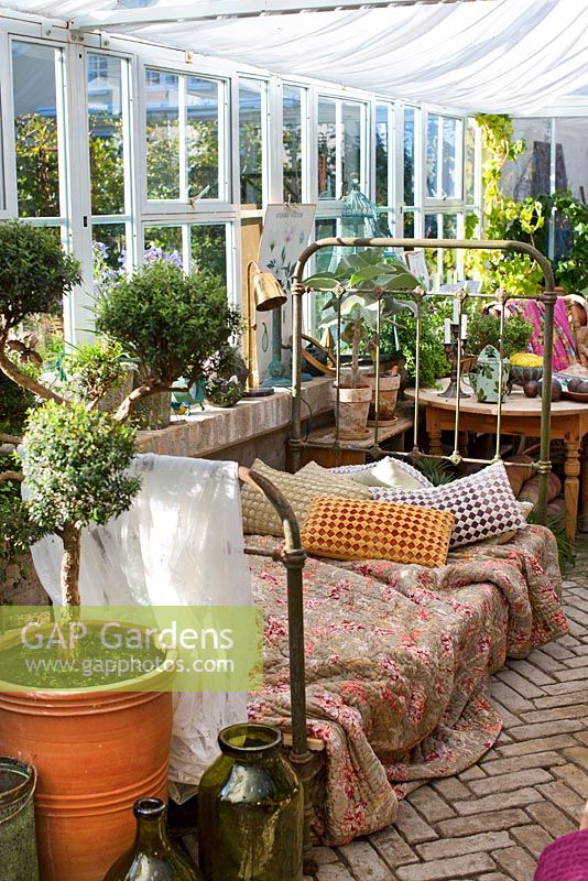 Greenhouse in garden, quilt and pillows on antique french iron bed, Myrtus in pots, brick floor 
