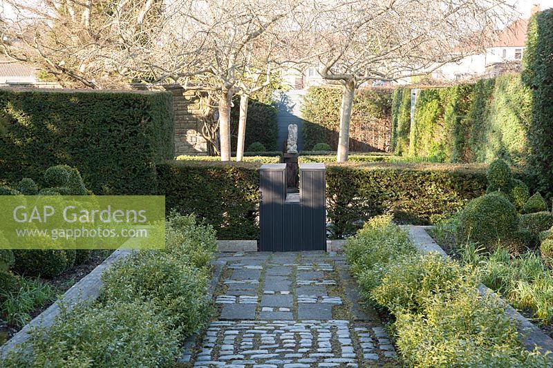 A path of paving slabs and stone setts is bordered by Christmas box, Sarcococca confusa, raised beds containing box topiary shapes and tall yew hedges. 