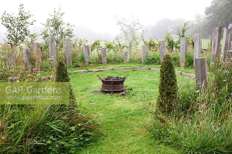 Wild meadow planted around a circular firepit on a misty morning, with oak obelisks and Buxus pyramids.