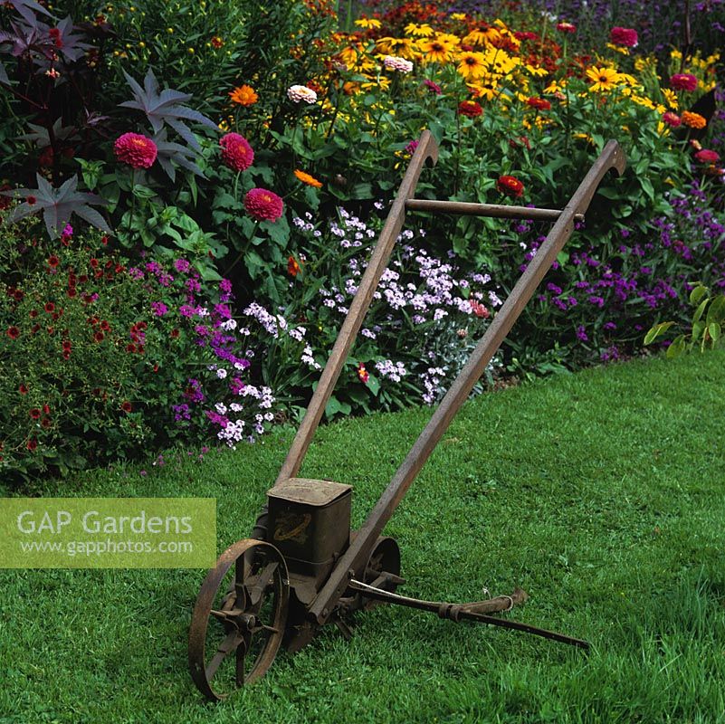 An old American Planet seeder rests on the grass by a summer bed of zinnia, verbena, helenium and gazania.