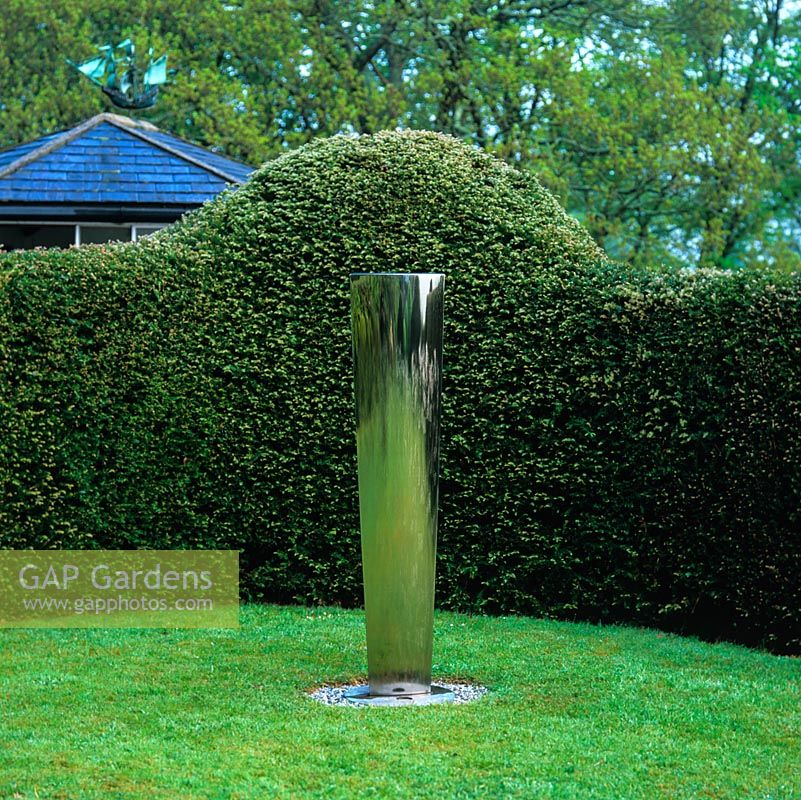 A stream of water runs down stainless steel sides of  'Water Tower' by Simon Percival, offset by curving yew hedges.