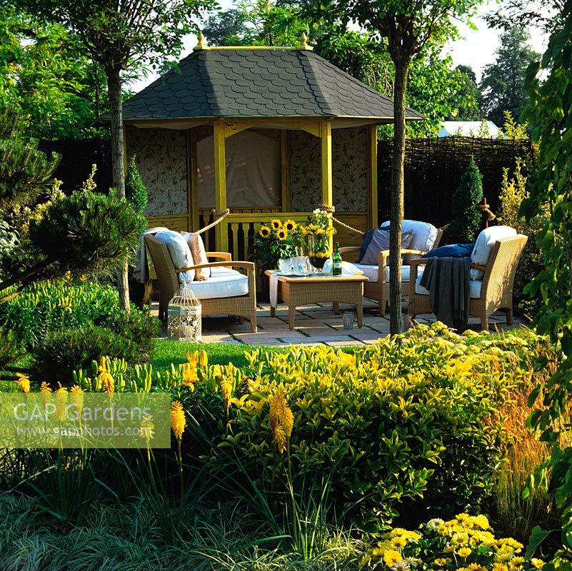 Tranquil retreat amidst gold and green plants - sunflower, kniphofia, lily, achillea, pine, box and elaeagnus. Summerhouse by patio with chairs and tables.
