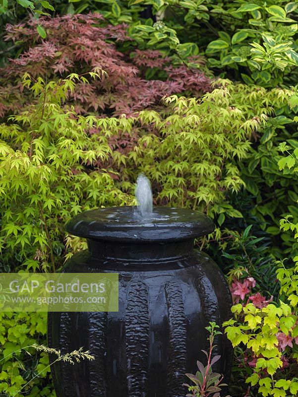 Huge ceramic urn has bubble fountain cascading water over its sides, against backdrop of maples.