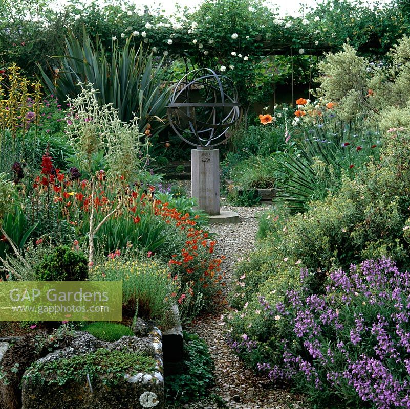 Gravel path in mediterranean style garden leads to armillary sphere and pergola. Beds of Salvia lavandulifolia, poppy, phlomis, yucca, bearded iris and helianthemum.