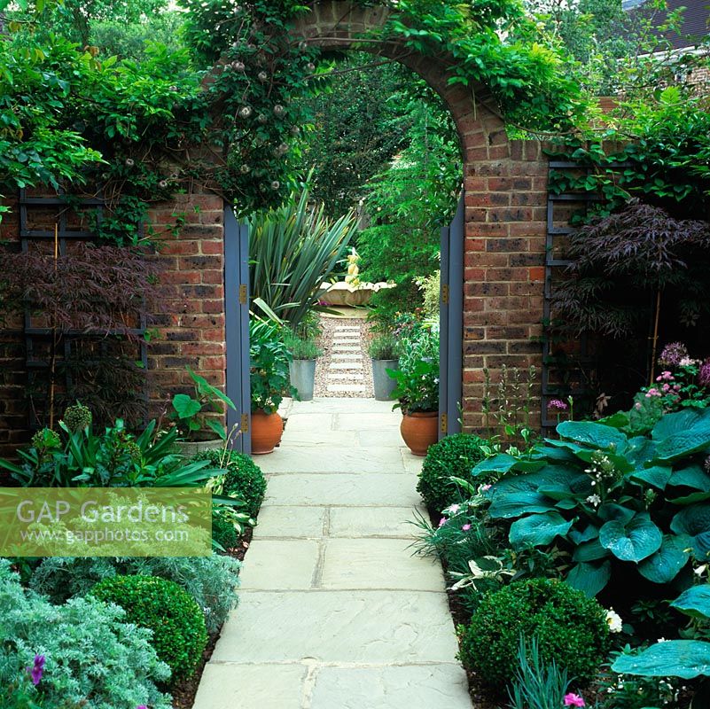 At side of house, paved path - edged in hosta, box, santolina and Allium cristophii - leads to arch framing view of path continuing to fountain. Creates illusion of greater space.