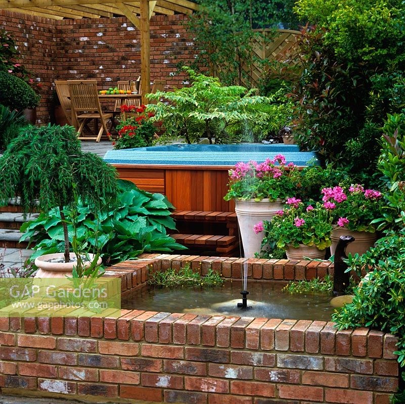 Outdoor hot tub is edged in geranium, hosta and photinia, partly concealed behind raised brick pool. Above, outdoor seating area beneath pergola.