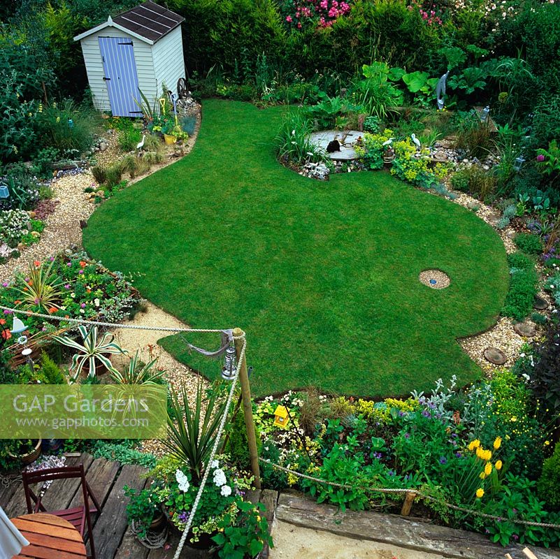Birds-eye view fish-shaped lawn enclosed by gravel path. Top: pond and deck edged in grasses. Shed. Below, sandpit and railway sleeper patio. Pots and raised woven hazel bed.