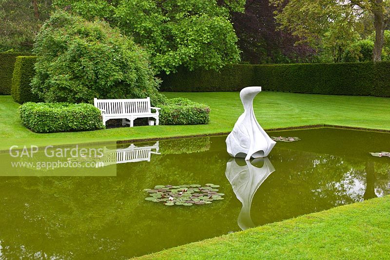 A tranquil water lily pond with a whale sculpture overlooked by a seat and surrounded by lawn, topiary and a Taxus hedge at Farleigh House, Hampshire