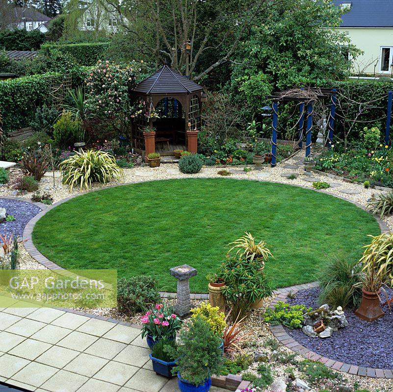 Town garden based on 3 linked circles - 2 slate beds by a brick-edged, circular lawn, edged by winding, gravel path. Gazebo. Pergola. Evergreen phormium, camellia, grasses.