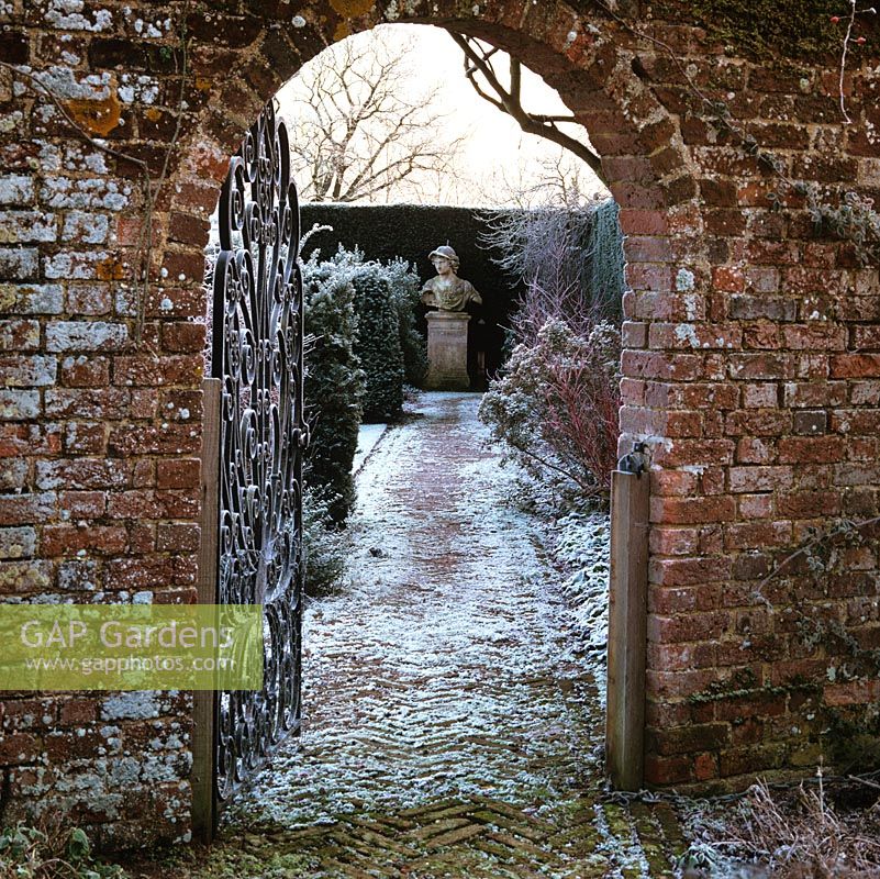 In ancient brick wall, iron gate opens to reveal frosty path leading to bust of Prince of Denmark set against yew hedges.