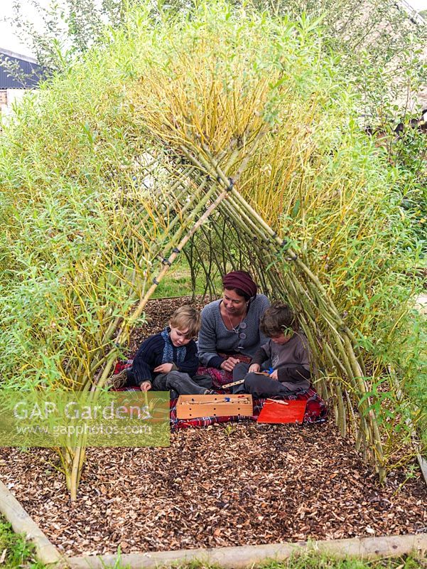 Brothers Jacob and Loui with their mother, Ali Rose, play on the floor in the shade of a tunnel  woven from living Salix viminalis - willow.