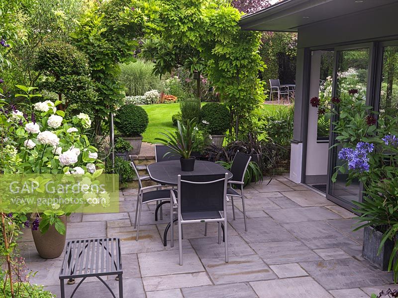 By kitchen, paved courtyard with pots of dahlia, agapanthus and white Hydrangea 'Black Steel Zambia'. Steps lead under wisteria arch, framing view of upper garden with miscanthus.
