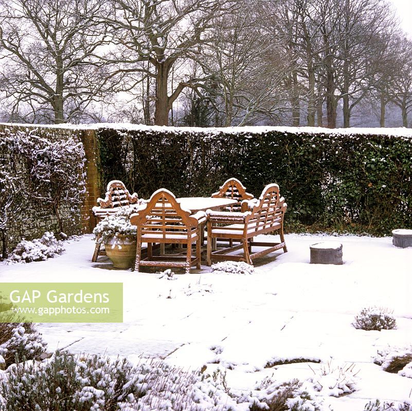 Lutyens style bench and table with dusting of snow on stone terrace sheltered by wall and hedge.