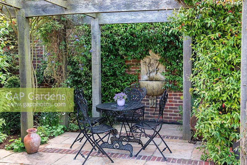 A stone and brick patio seating area covered by a wooden pergola. On the back wall a ceramic water feature from Lucy Smith Garden Sculptures