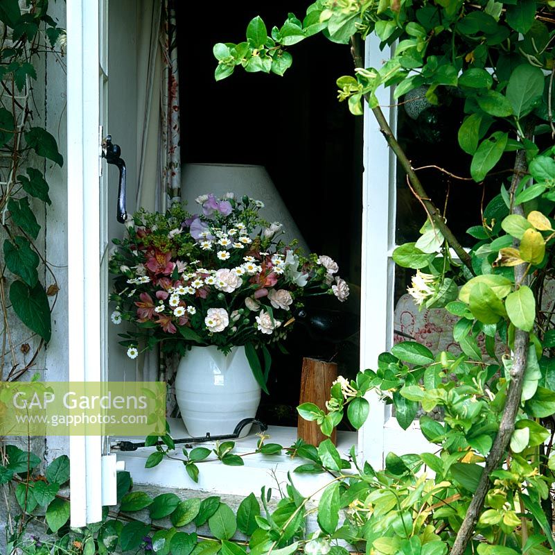 Wooden bird ornament sits on cottage windowsill beside vase of flowers.