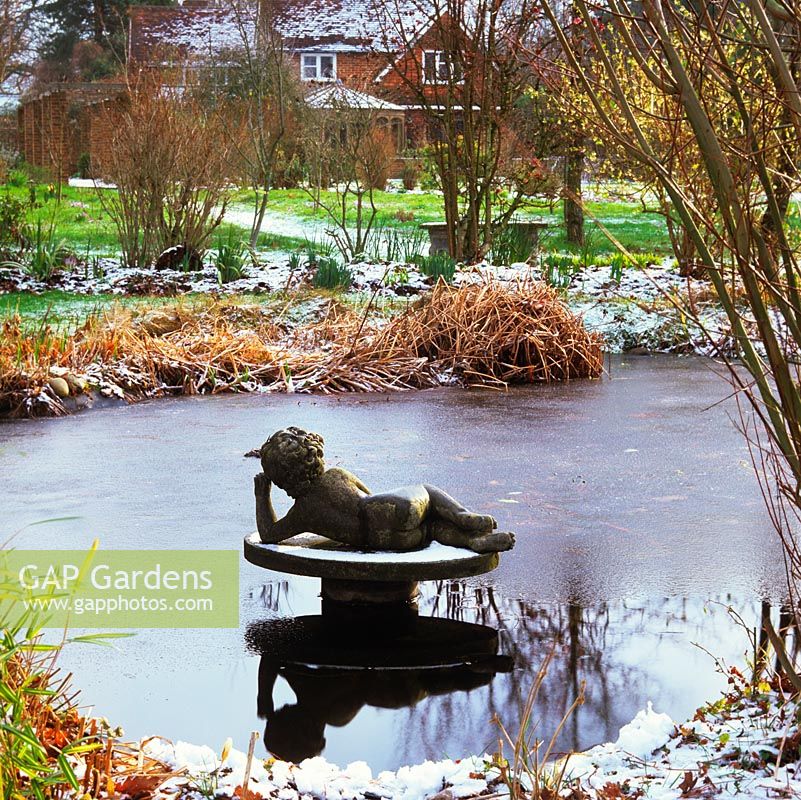 A frozen wildlife pond, a reclining cherub fountain at its centre, reflects naked trees. Fringed in snow encrusted grasses.