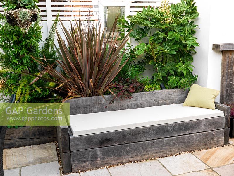 Outdoor room with built-in seating, mirror and raised bed of phormium, solanum, fatsia, fern. Mirror behind foliage.