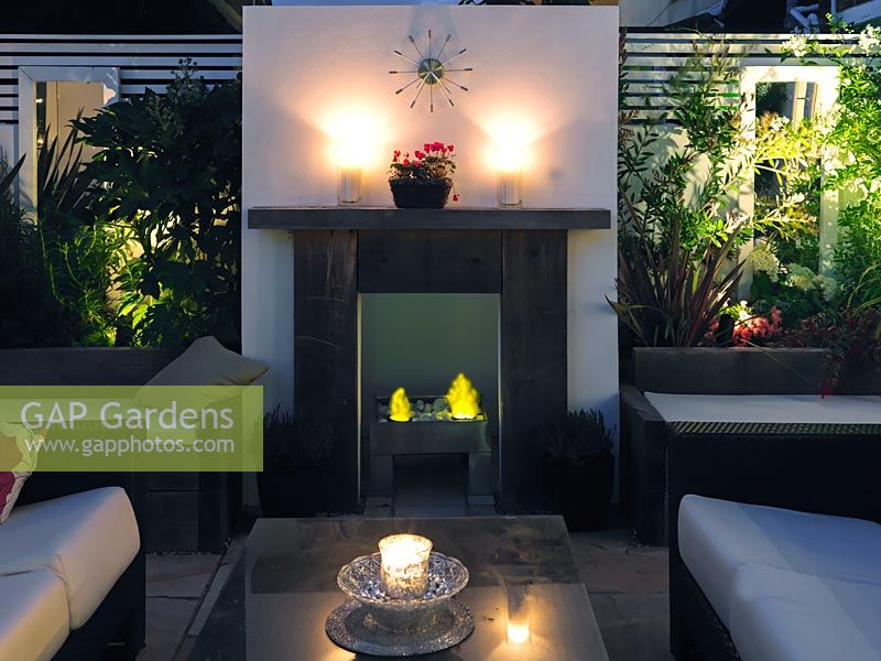 Outdoor room at night with sofas by gas effect fire and fireplace. Backlit raised beds to each side, behind built-in seating.