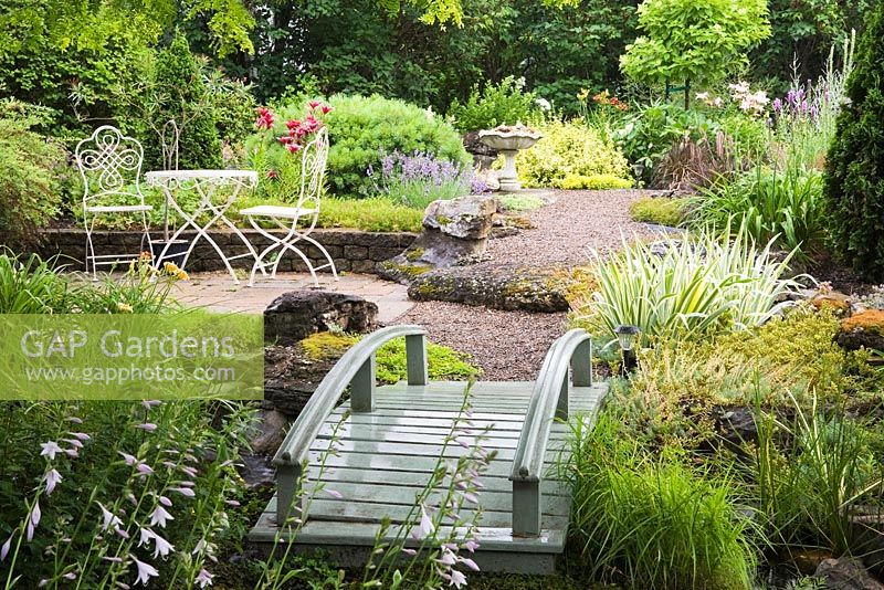 Grey painted wooden footbridge bordered by Acorus gramineus 'Variegatus - Ornamental Grass plants leading to a white cast-iron metal table and chairs in backyard country garden in summer, Quebec, Canada