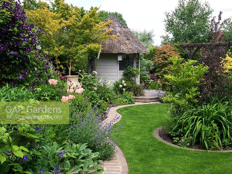 A shaped grass path leads through mature mixed borders to a thatched summer house.